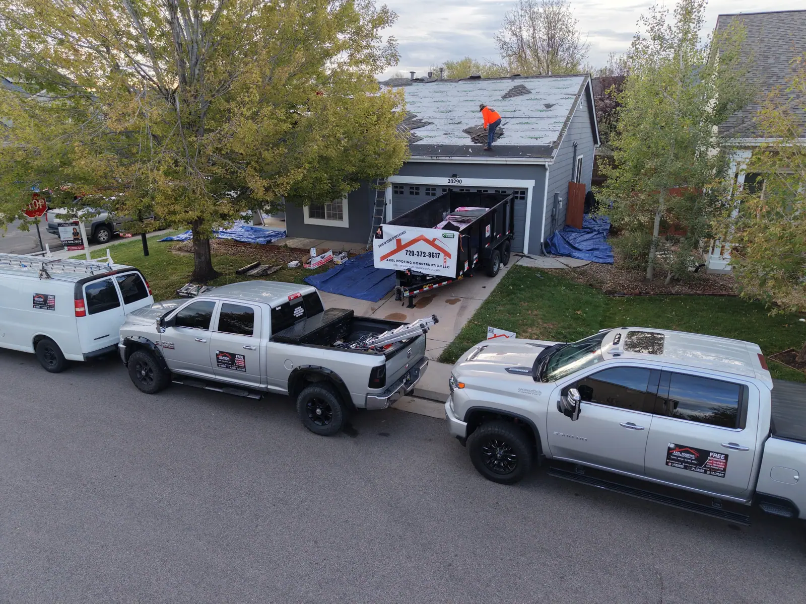 AXEL Roofing trucks and branded dump trailer parked at a home while a roofer tears off the old shingles